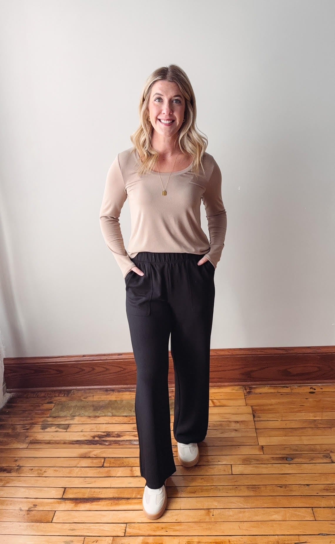 Woman wearing a beige top and black pants standing on a wooden floor with a white wall background