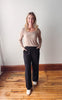 Woman wearing a beige top and black pants standing on a wooden floor with a white wall background