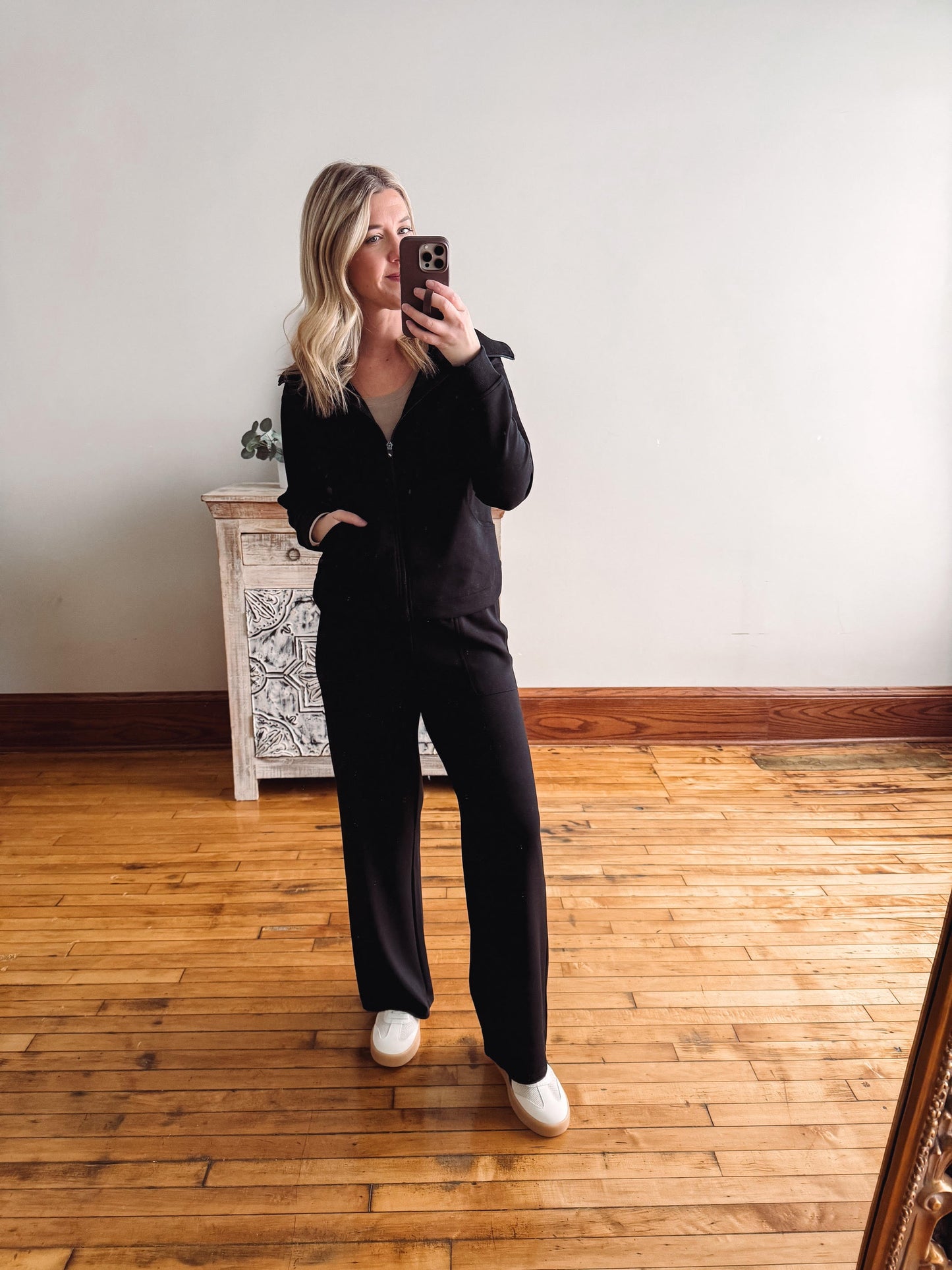 Person wearing a black outfit taking a mirror selfie on a wooden floor.