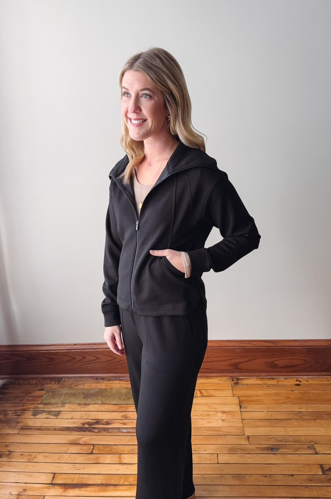 Woman wearing a black hoodie and pants standing on a wooden floor with a white wall background