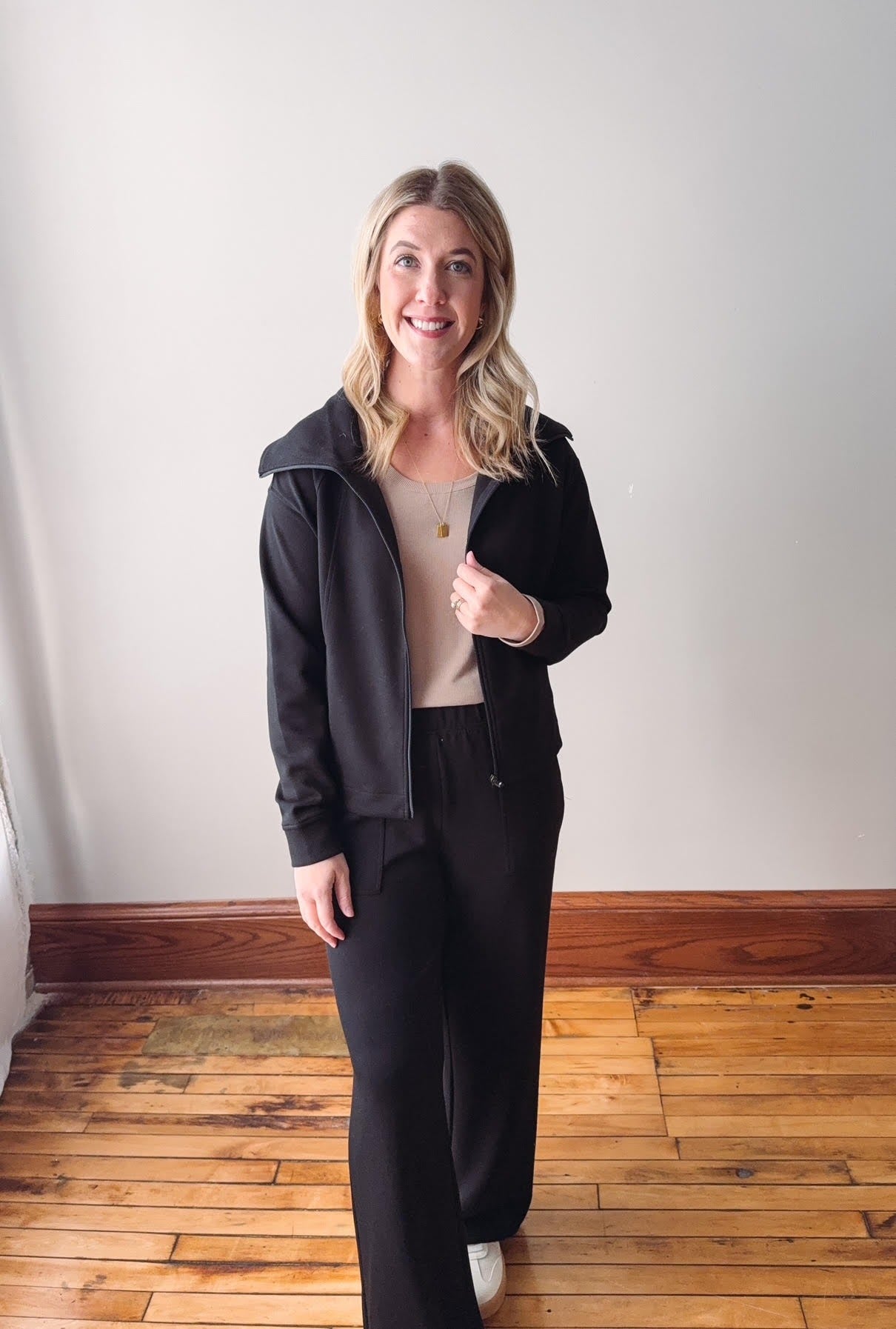 Woman wearing a black outfit standing on a wooden floor with a white wall background