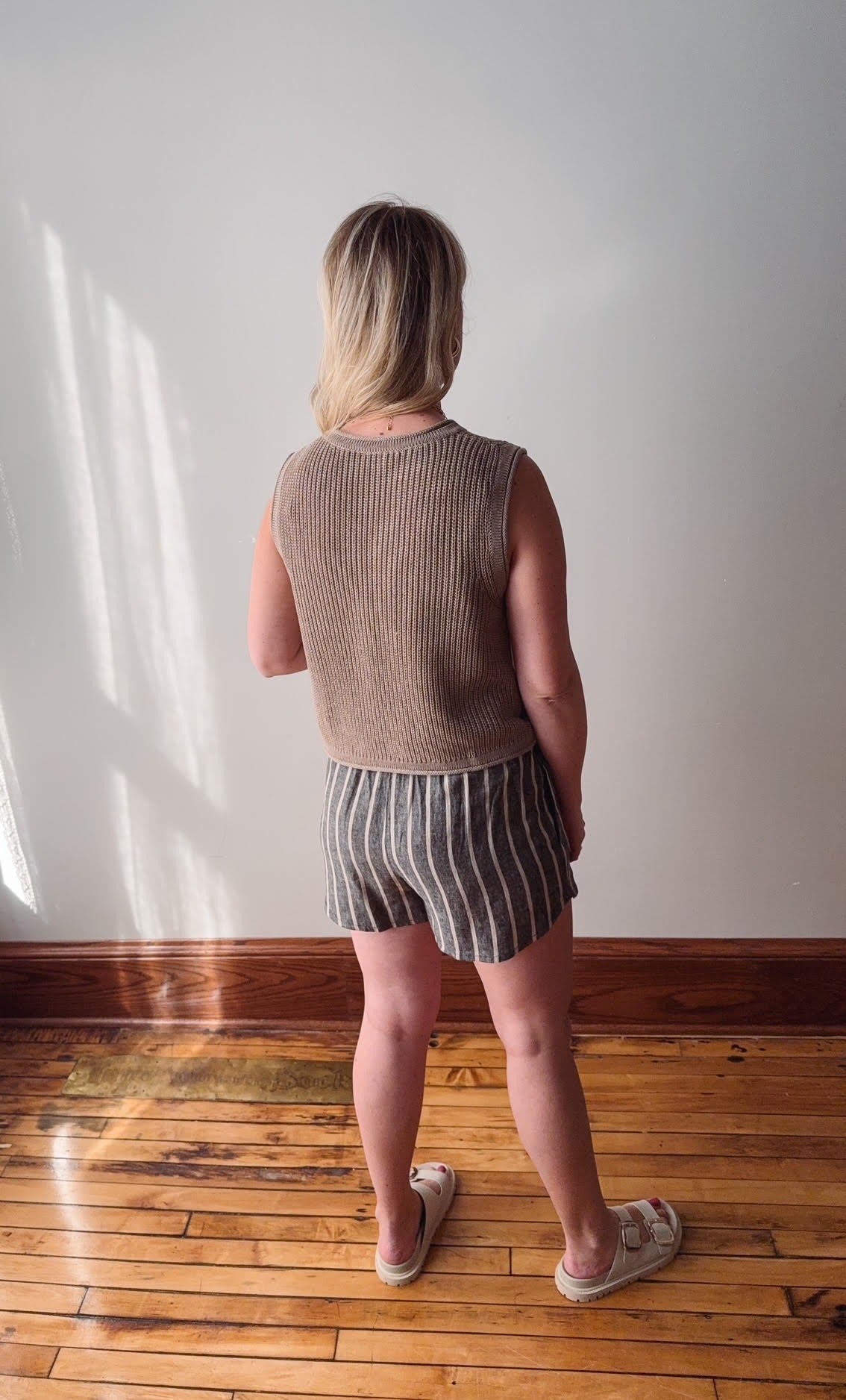 Person wearing a sleeveless brown top and striped shorts standing in a room with wooden flooring and white walls.