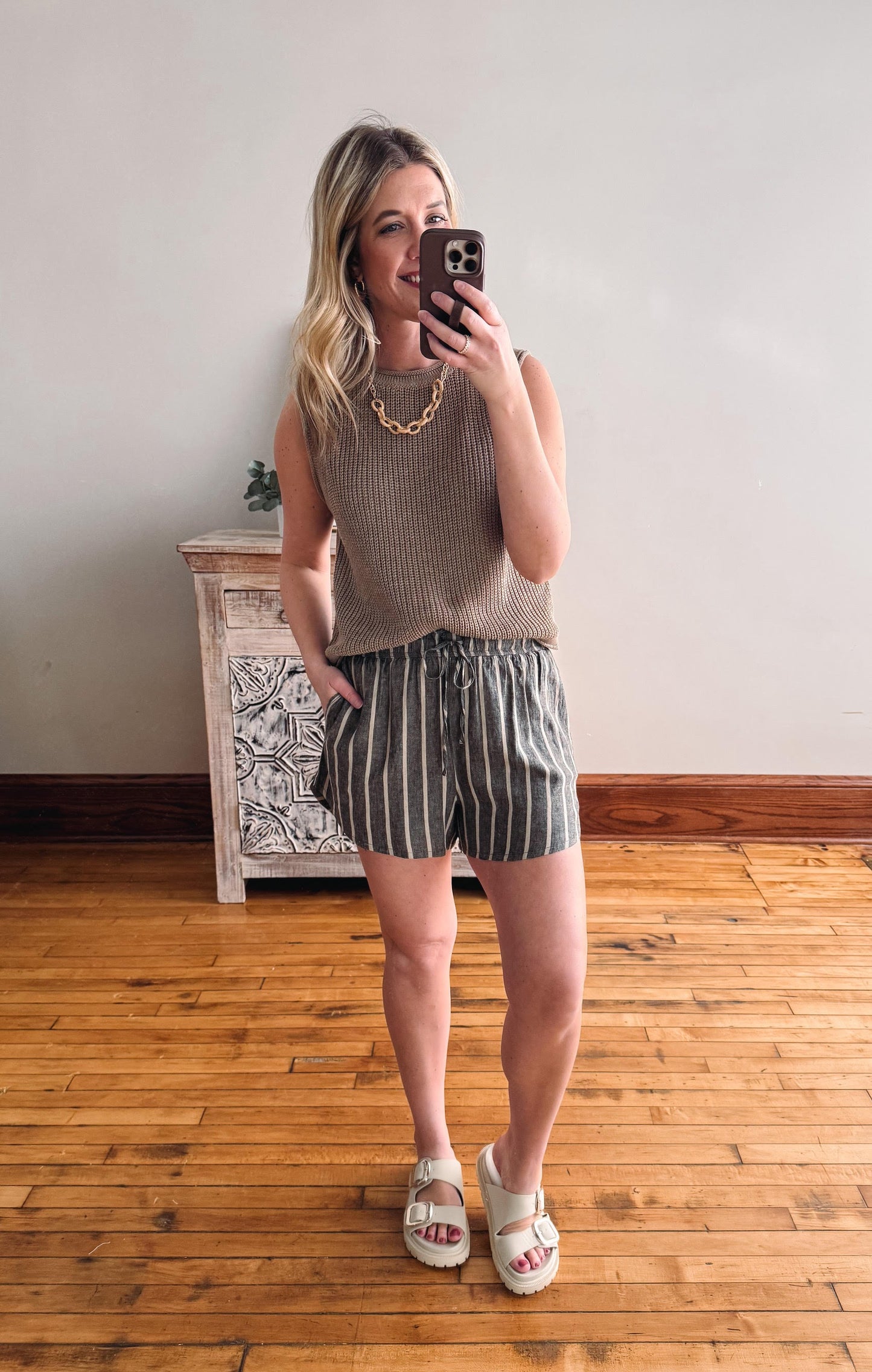 Woman taking a mirror selfie wearing a sleeveless top and striped shorts in a room with wooden flooring.