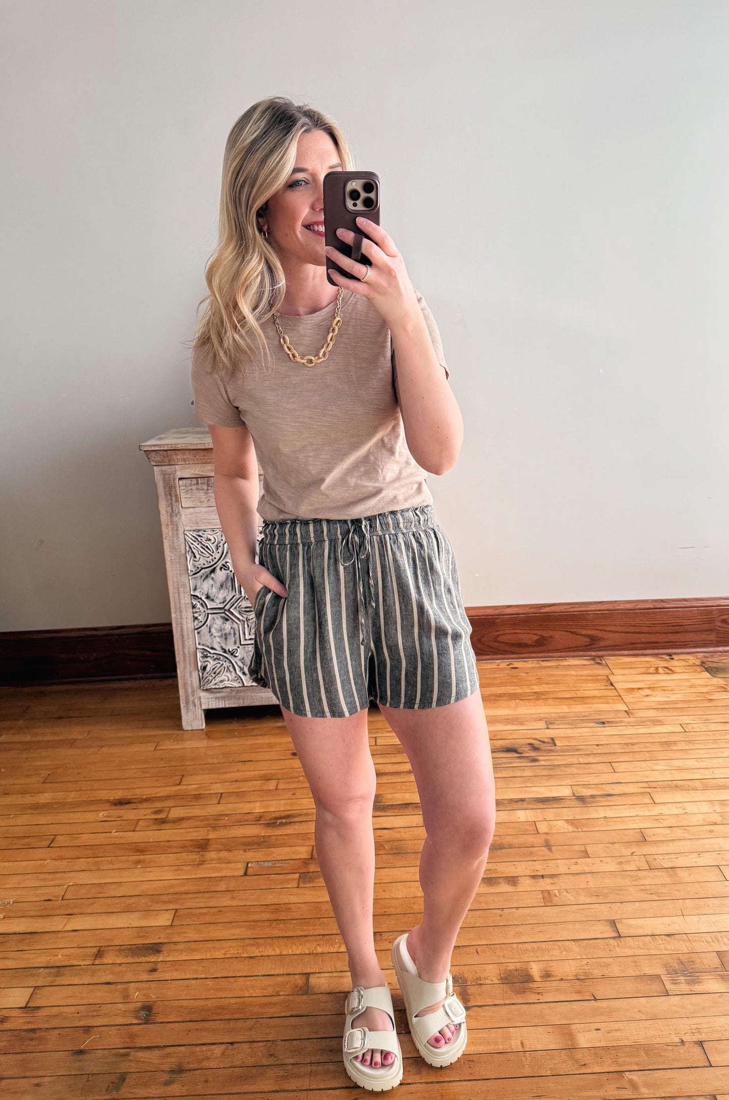 Woman taking a mirror selfie wearing a beige top and striped shorts in a room with wooden flooring.