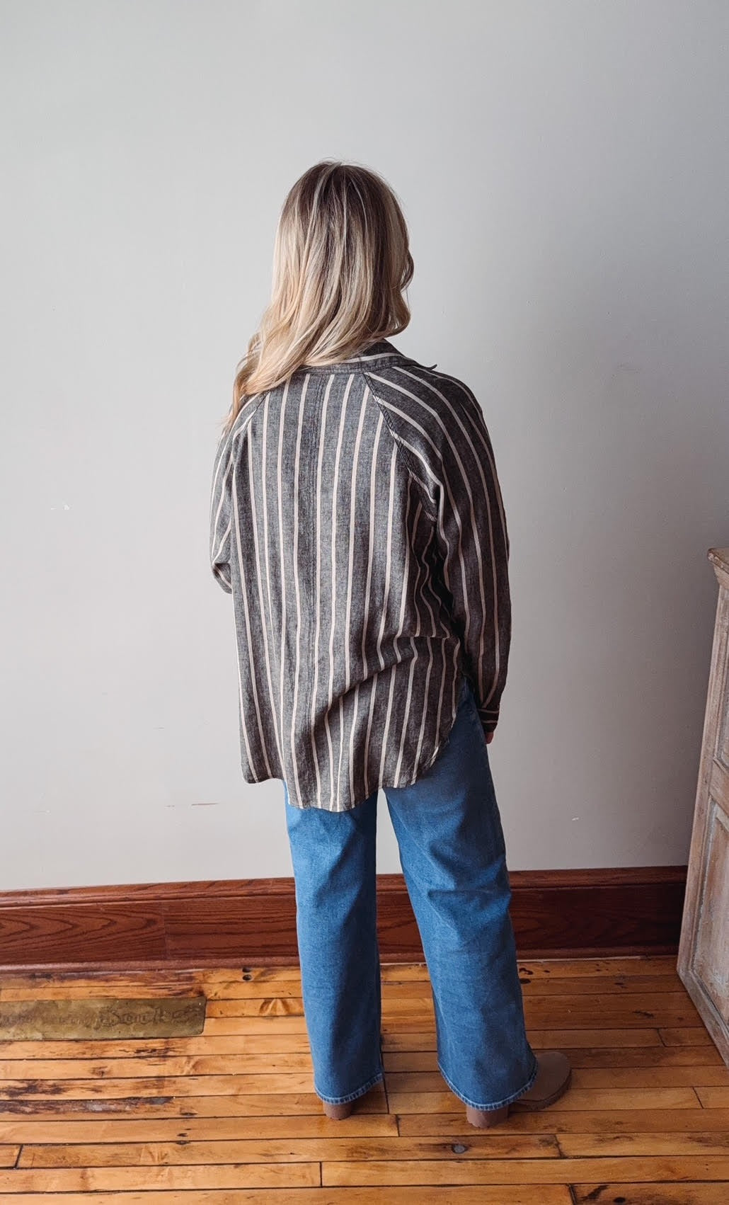 Person wearing a striped shirt and blue jeans standing in a room with wooden flooring and a white wall.