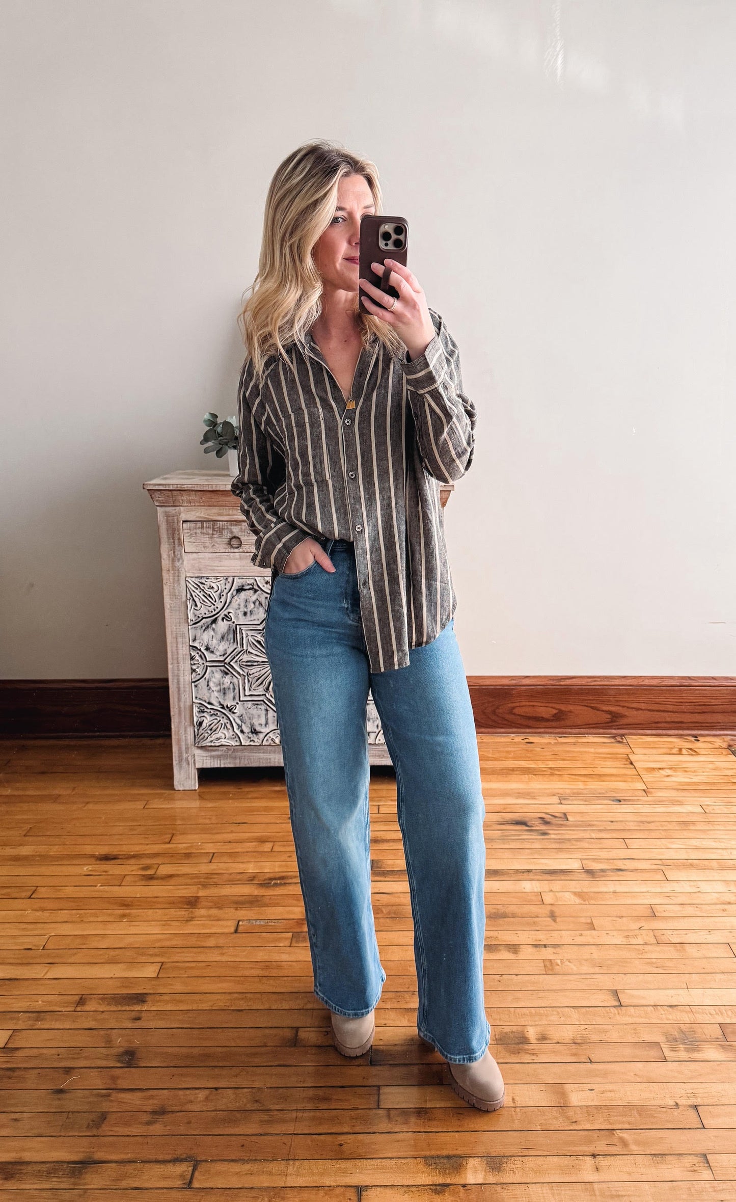 Woman taking a mirror selfie wearing a striped shirt and jeans in a room with wooden flooring and a decorative cabinet.