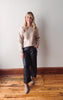 Woman wearing a beige sweater and black pants standing against a plain wall.