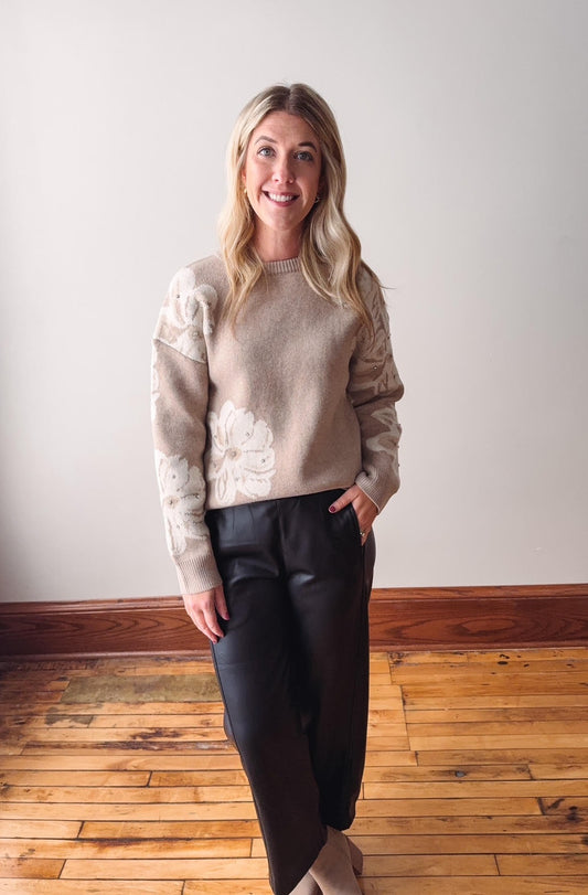 Woman wearing a beige sweater with floral details and black pants standing on a wooden floor.