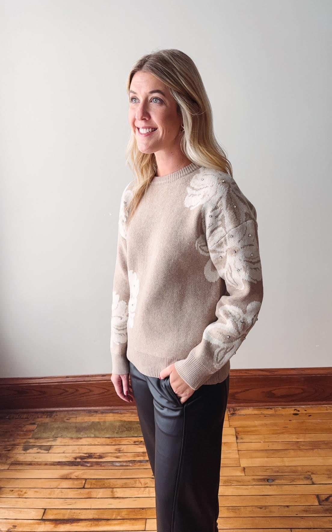 Woman wearing a beige sweater with floral patterns in a room with wooden flooring and a white wall.