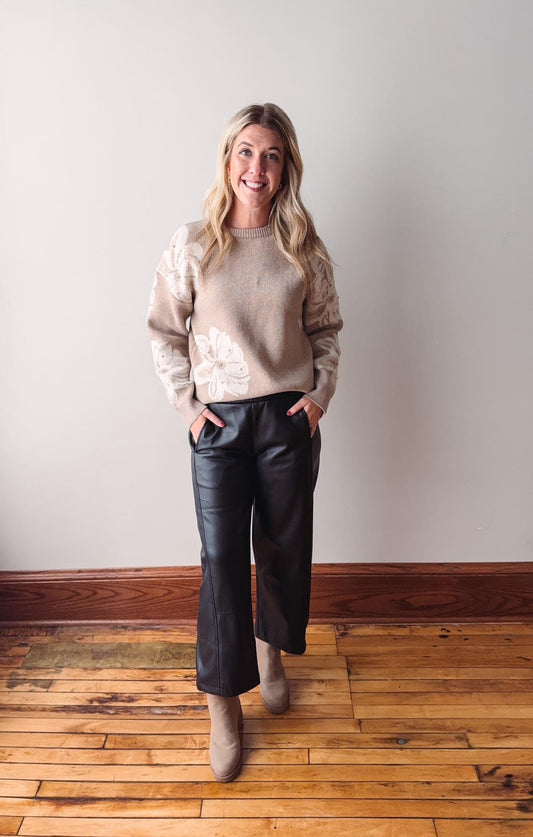 Woman wearing a beige sweater and black leather pants standing on a wooden floor against a white wall.