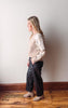 Woman standing against a white wall wearing a beige sweater with lace details and dark pants.