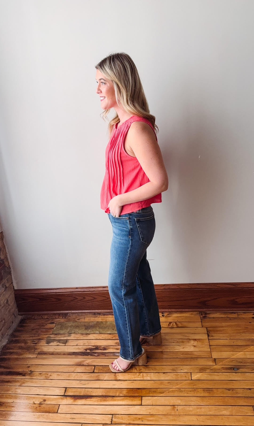 Woman wearing a pink top and blue jeans standing against a white wall on a wooden floor.