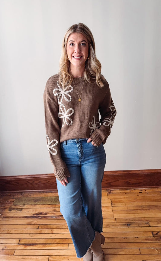 Woman wearing a brown sweater with white floral patterns and blue jeans indoors.