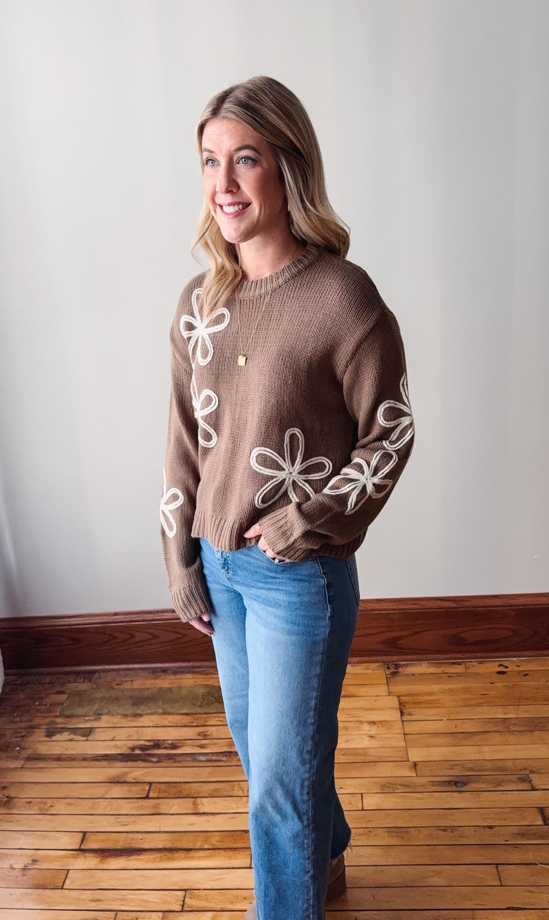 Woman wearing a brown sweater with white floral patterns and blue jeans standing on a wooden floor.
