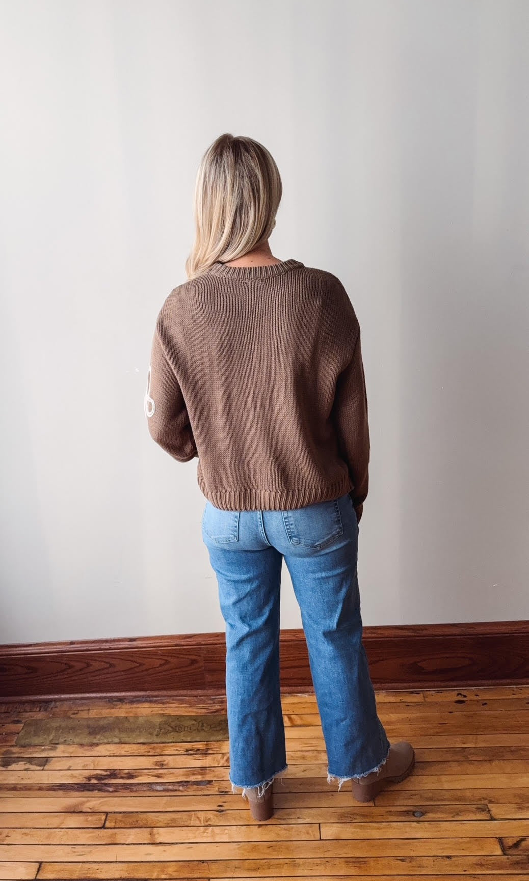 Person wearing a brown sweater and blue jeans standing against a plain wall.
