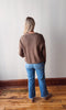 Person wearing a brown sweater and blue jeans standing against a plain wall.