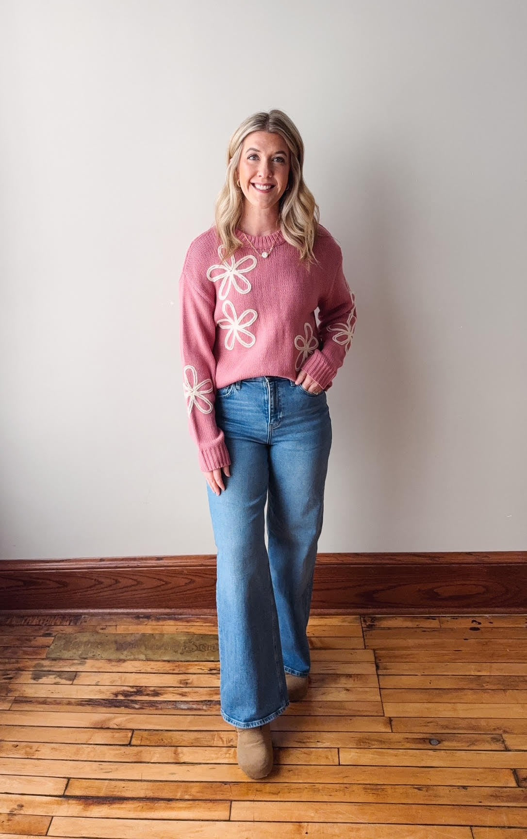 Woman wearing a pink sweater with white floral designs and blue jeans standing on a wooden floor.