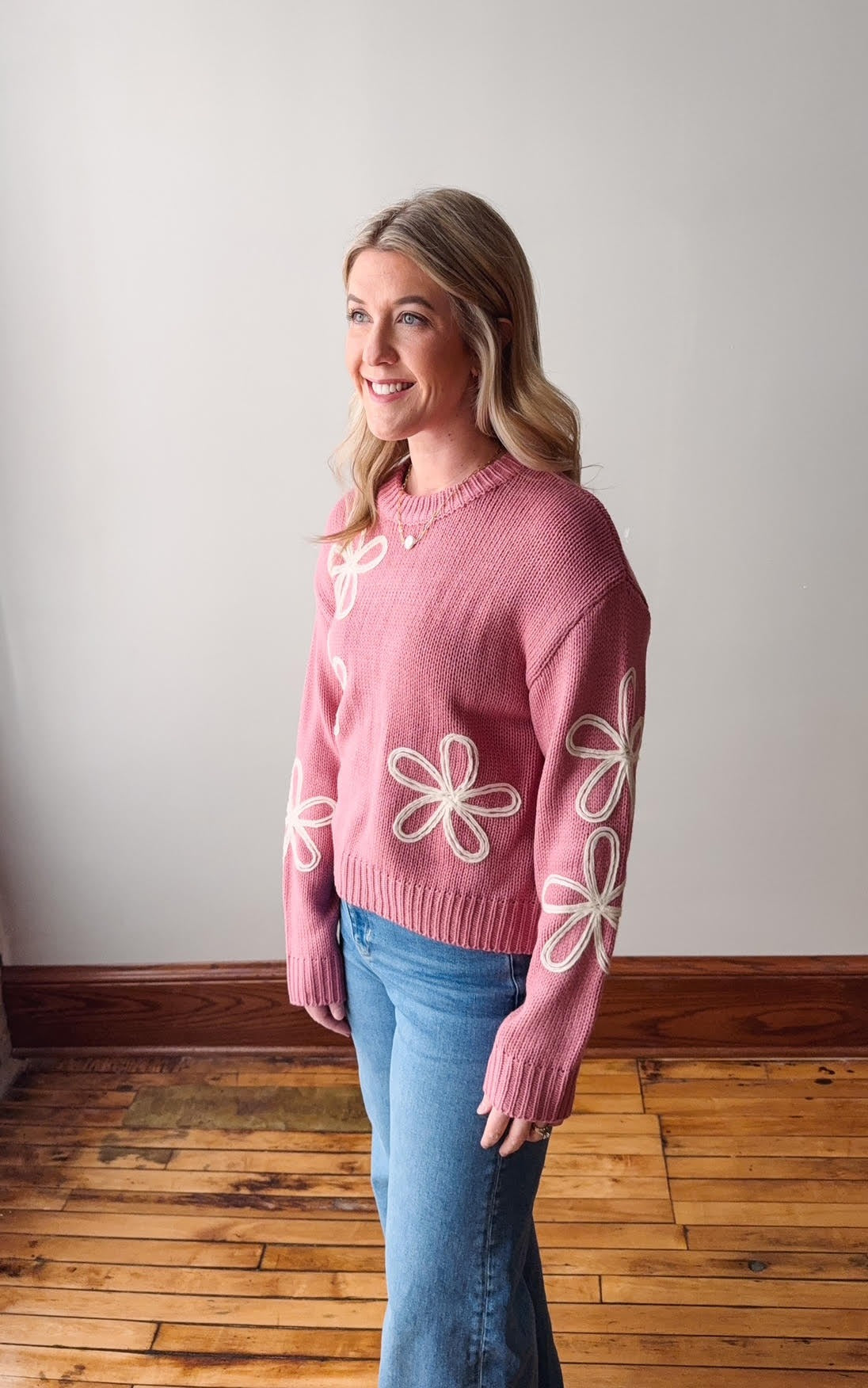 Woman wearing a pink sweater with white floral patterns in a room with wooden flooring.
