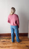 Person wearing a pink sweater and blue jeans standing on a wooden floor against a white wall.
