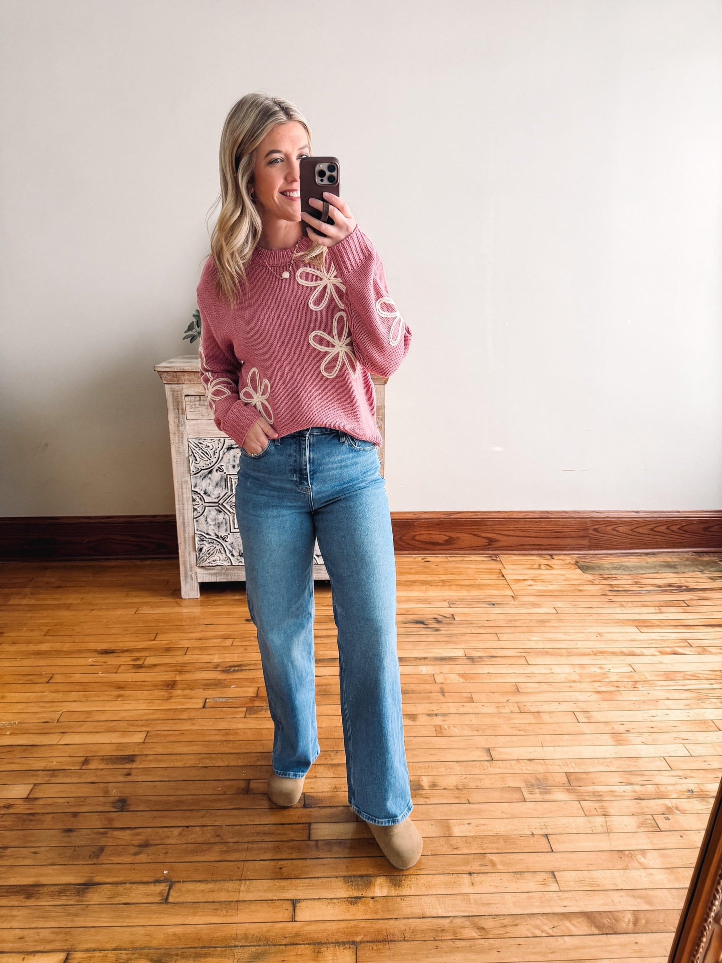 Woman taking a mirror selfie wearing a pink sweater with floral patterns and blue jeans in a room with wooden flooring.