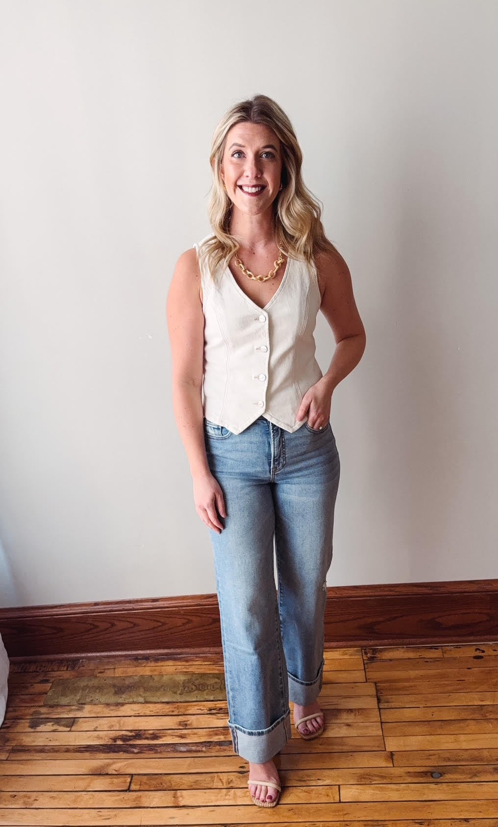 Woman wearing a white sleeveless top and blue jeans standing on a wooden floor against a white wall.