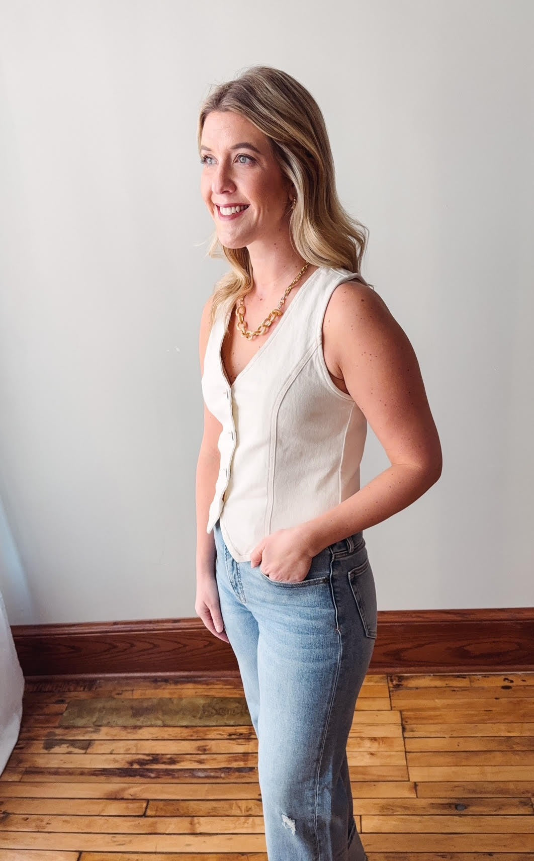 Woman wearing a white sleeveless top and light blue jeans standing in a room with wooden flooring.