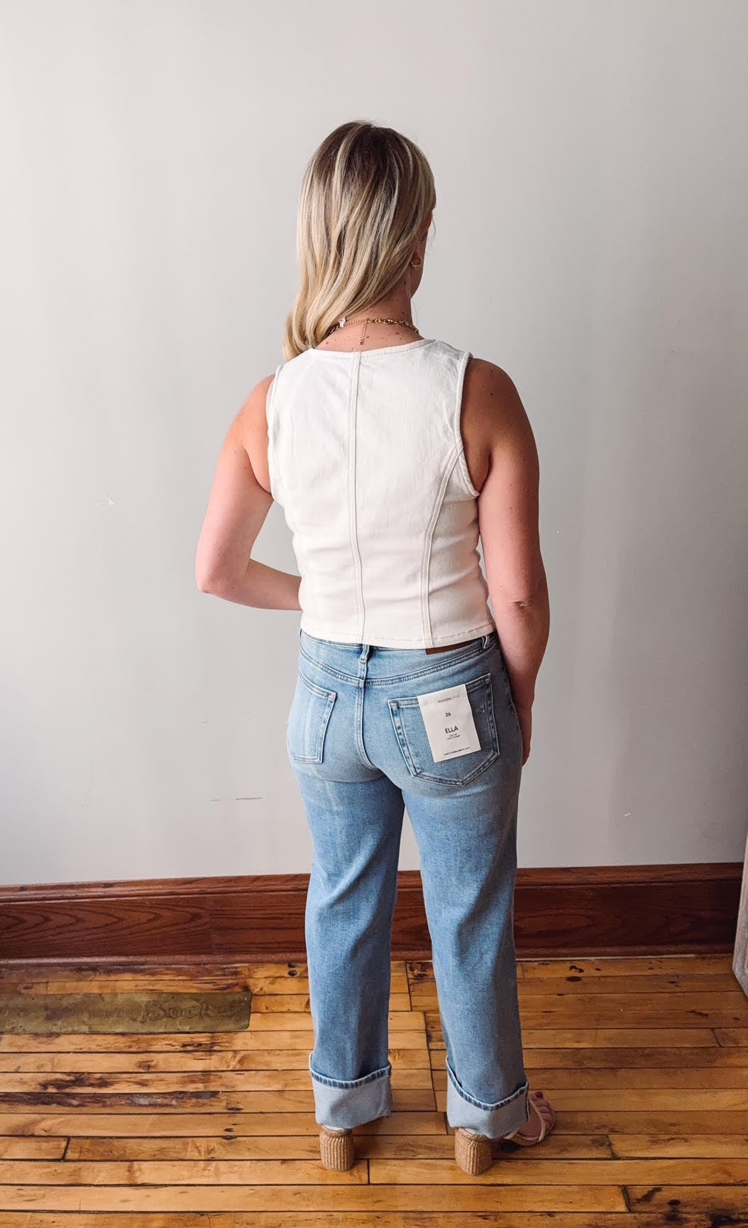 Person wearing a white sleeveless top and blue jeans standing on a wooden floor with a plain wall background.
