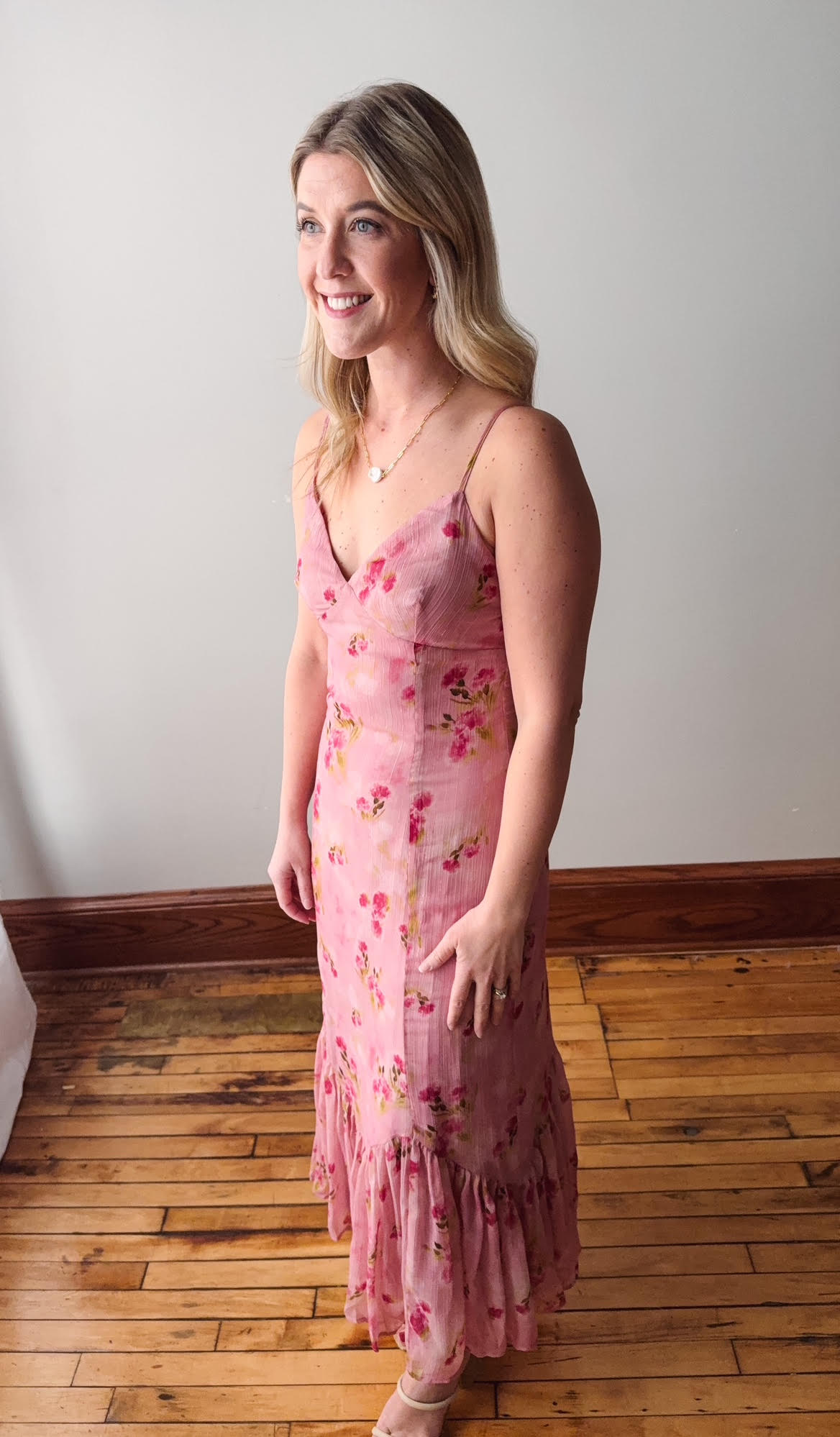 Woman wearing a pink floral dress standing on a wooden floor with a white wall background