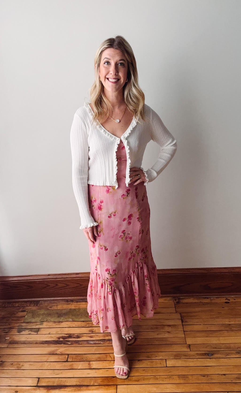 Woman wearing a white blouse and pink floral skirt standing on a wooden floor against a white wall.