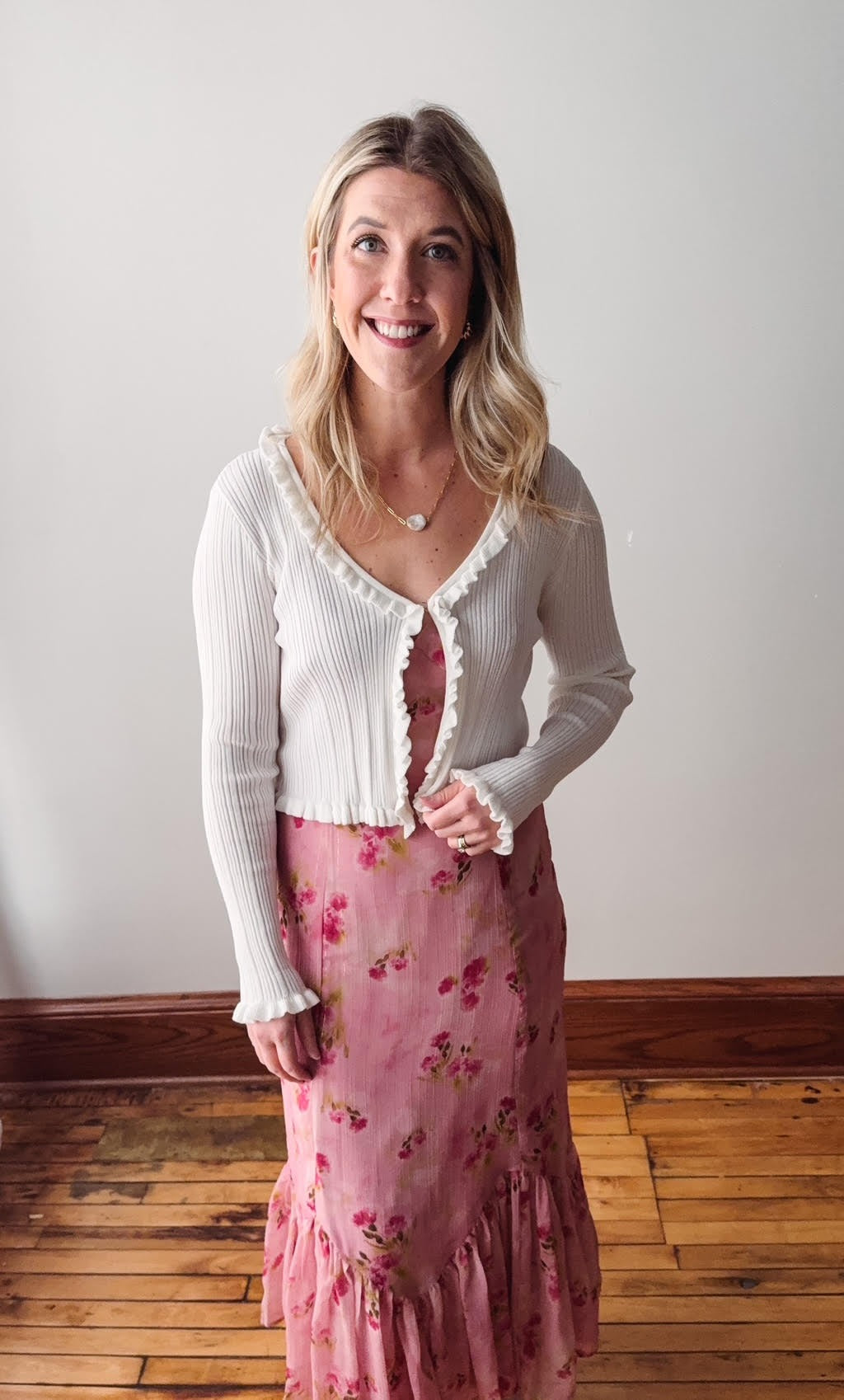 Woman wearing a white cardigan and pink floral skirt standing against a plain wall.