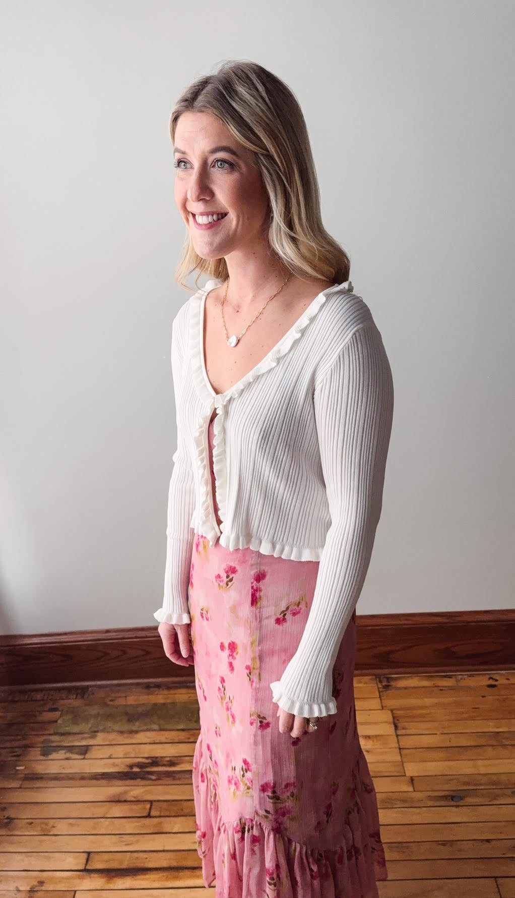 Woman wearing a white cardigan and pink floral skirt standing against a plain wall.