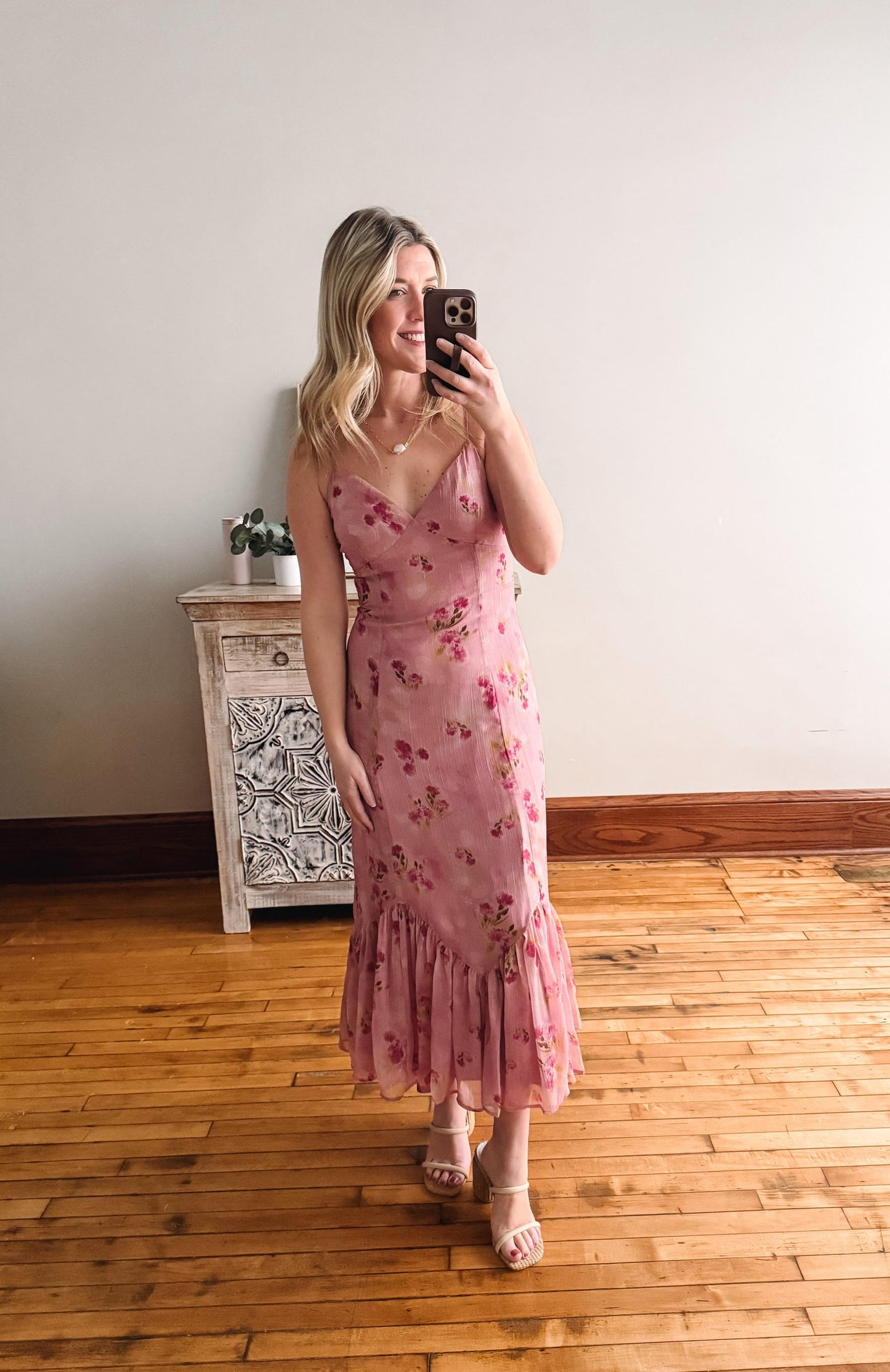 Woman taking a mirror selfie wearing a pink floral dress in a room with wooden flooring and a decorative cabinet.