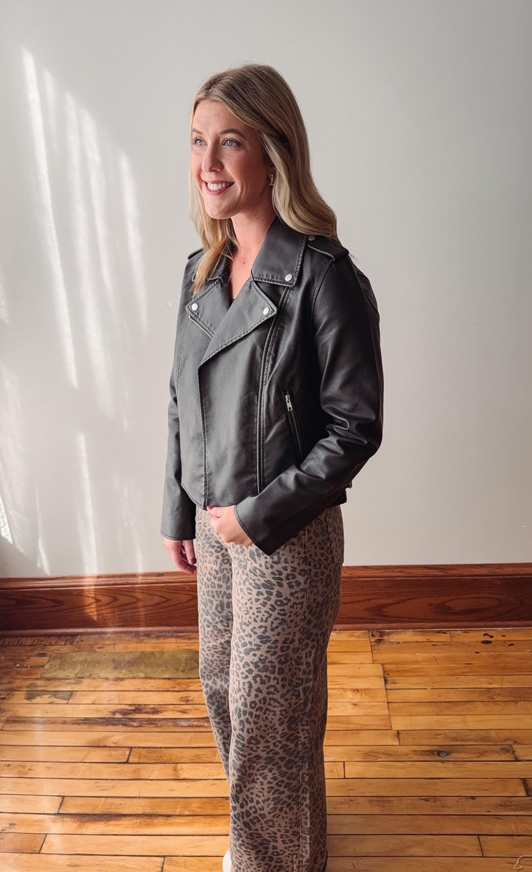 Woman wearing a black leather jacket and patterned pants standing in a room with wooden flooring.