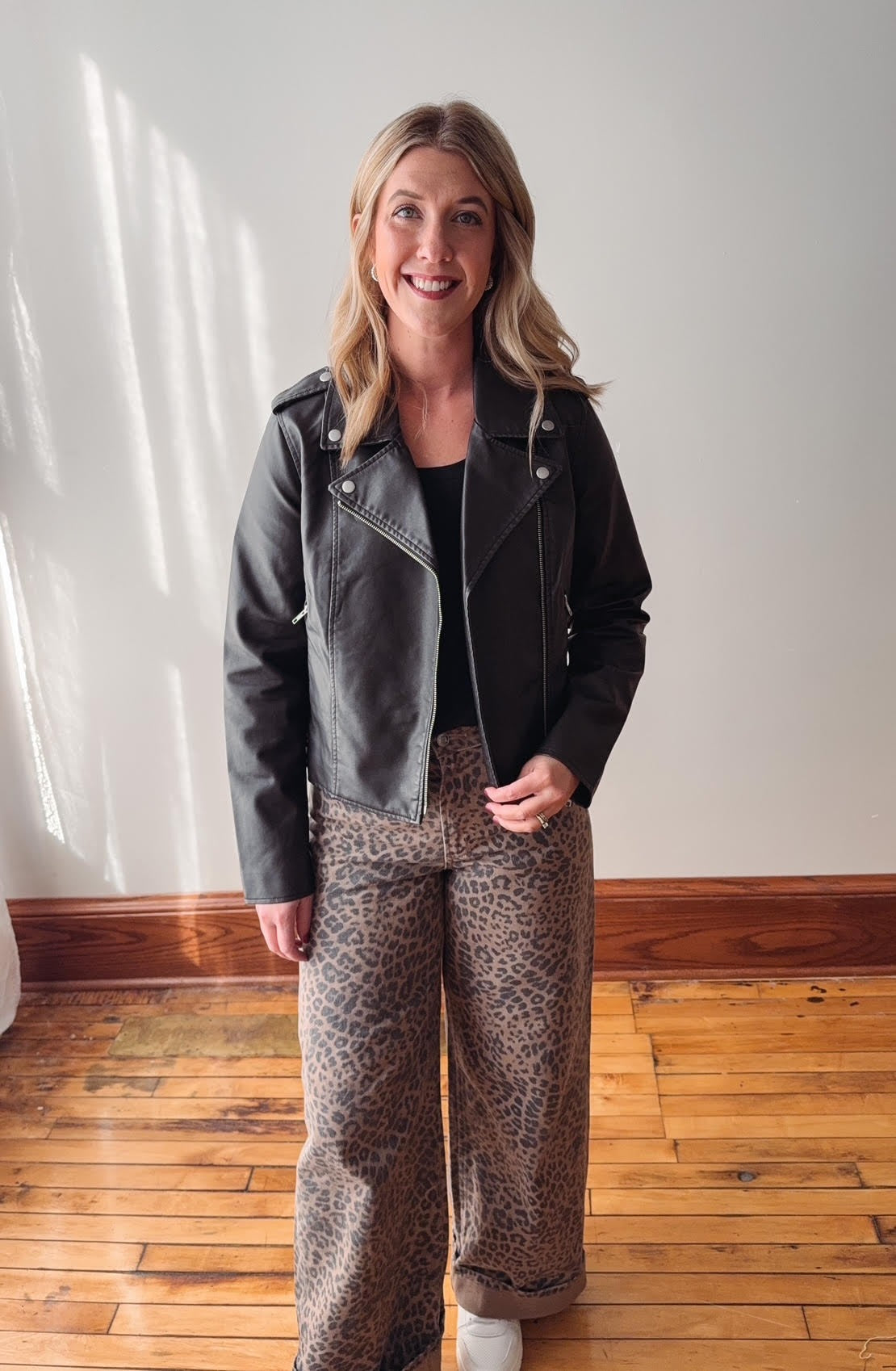 Woman wearing a black leather jacket and patterned pants standing on a wooden floor.