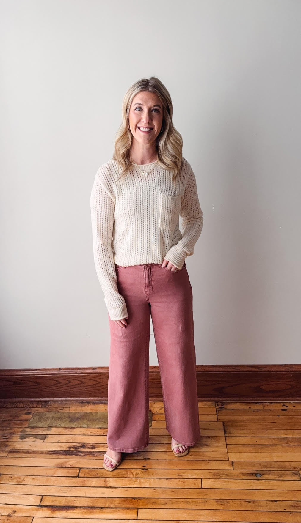 Woman wearing a white long-sleeve top and pink pants standing against a plain wall.