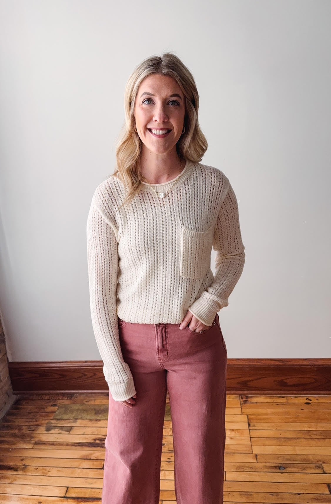 Woman wearing a cream sweater and pink pants standing against a white wall.