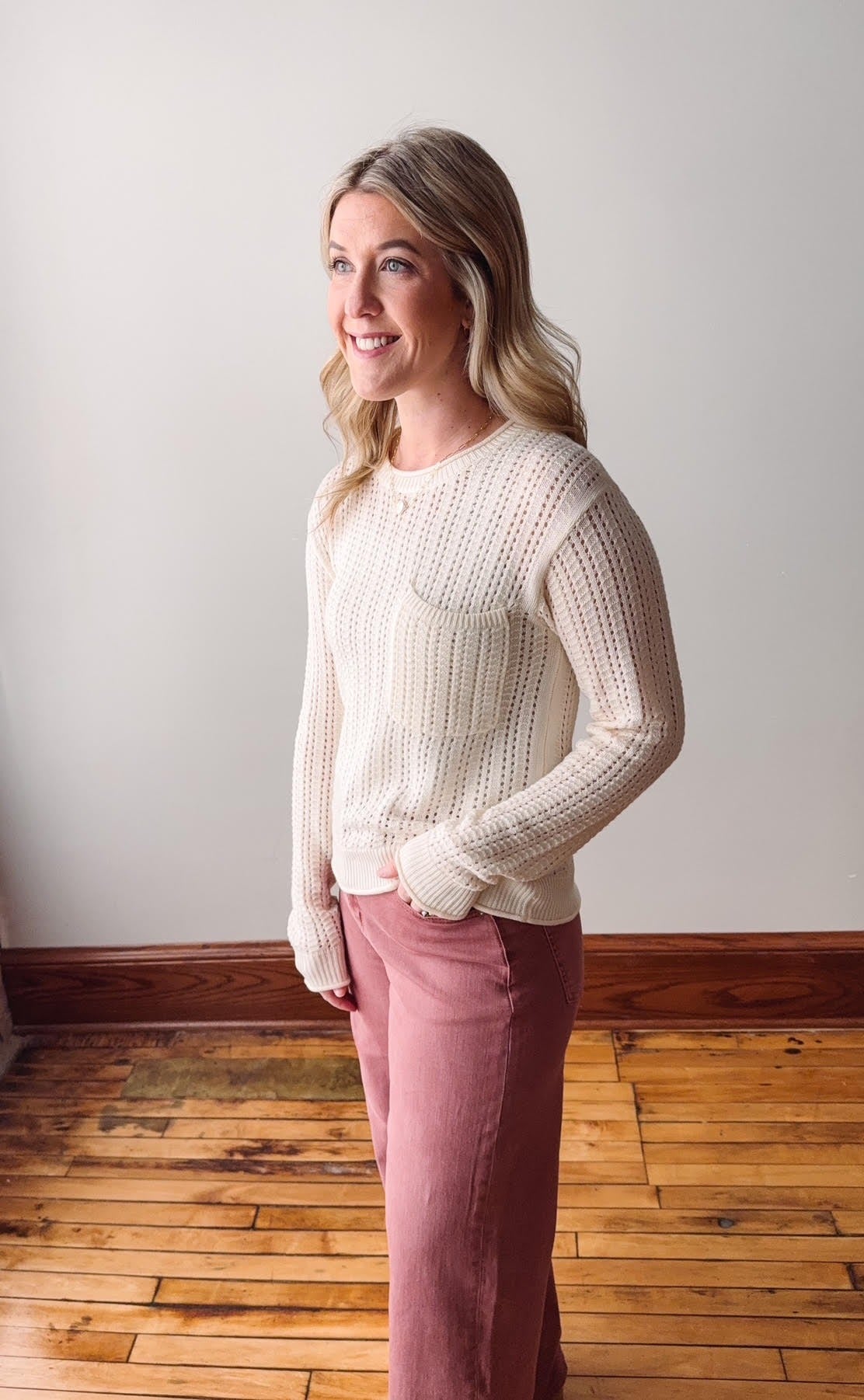 Woman wearing a cream sweater and pink pants standing against a white wall.