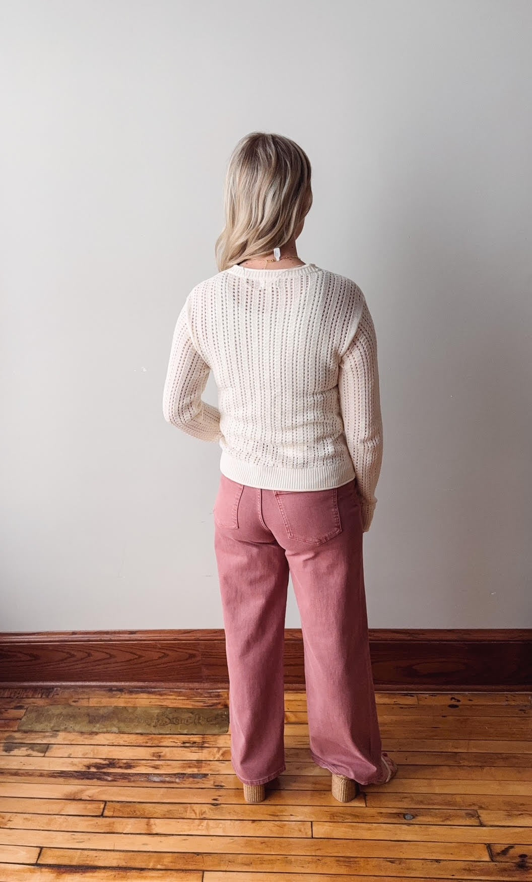 Person wearing a white sweater and pink pants standing on a wooden floor against a white wall.