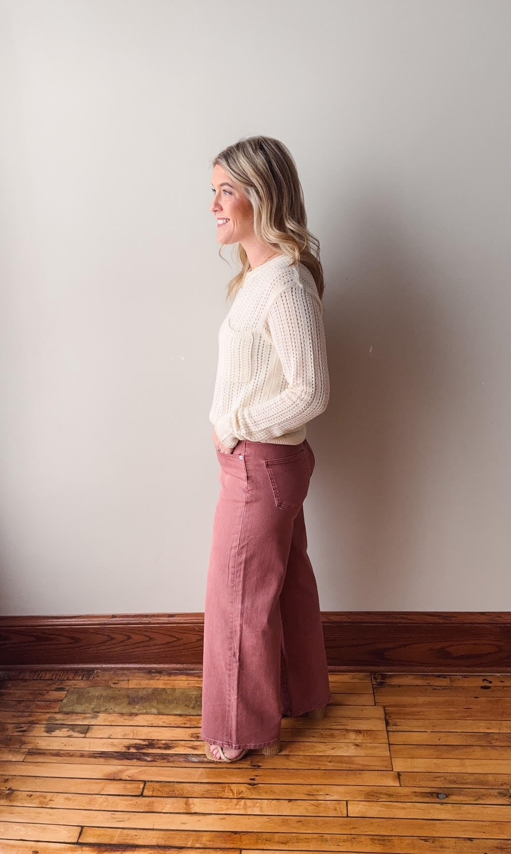 Woman wearing a white blouse and pink pants standing against a plain wall.