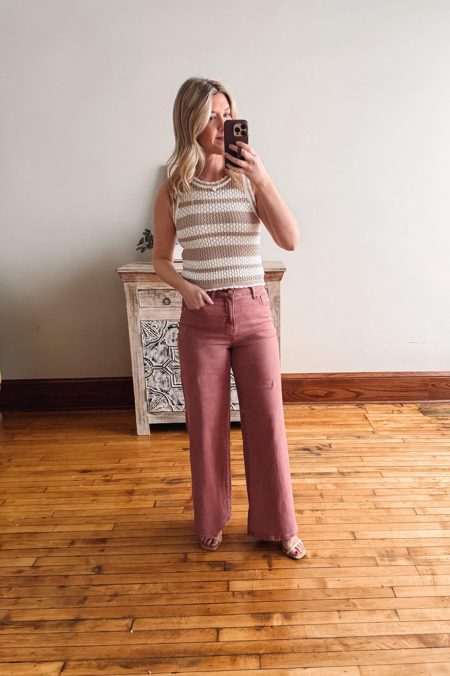 Woman taking a mirror selfie wearing a striped sleeveless top and pink pants in a room with wooden flooring.