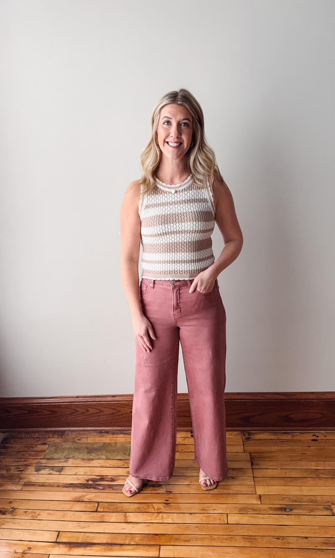 Woman wearing a striped sleeveless top and pink pants standing in a room with wooden flooring and a white wall.