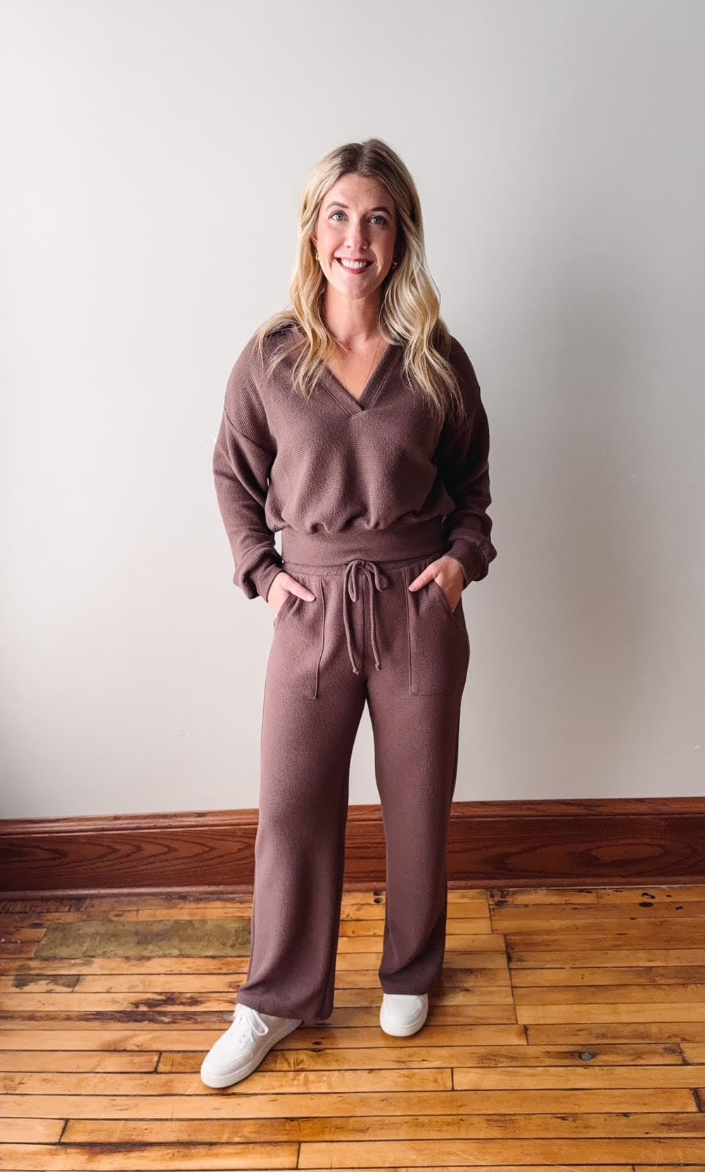 Woman wearing a brown outfit standing on a wooden floor with a white wall background
