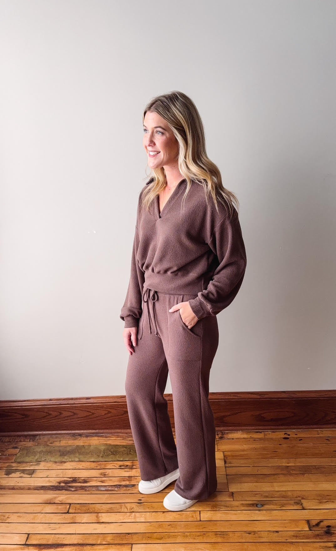Woman wearing a brown outfit standing on a wooden floor against a white wall