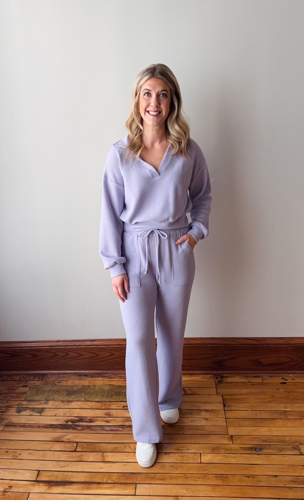 Woman wearing a light purple outfit standing on a wooden floor with a white wall background