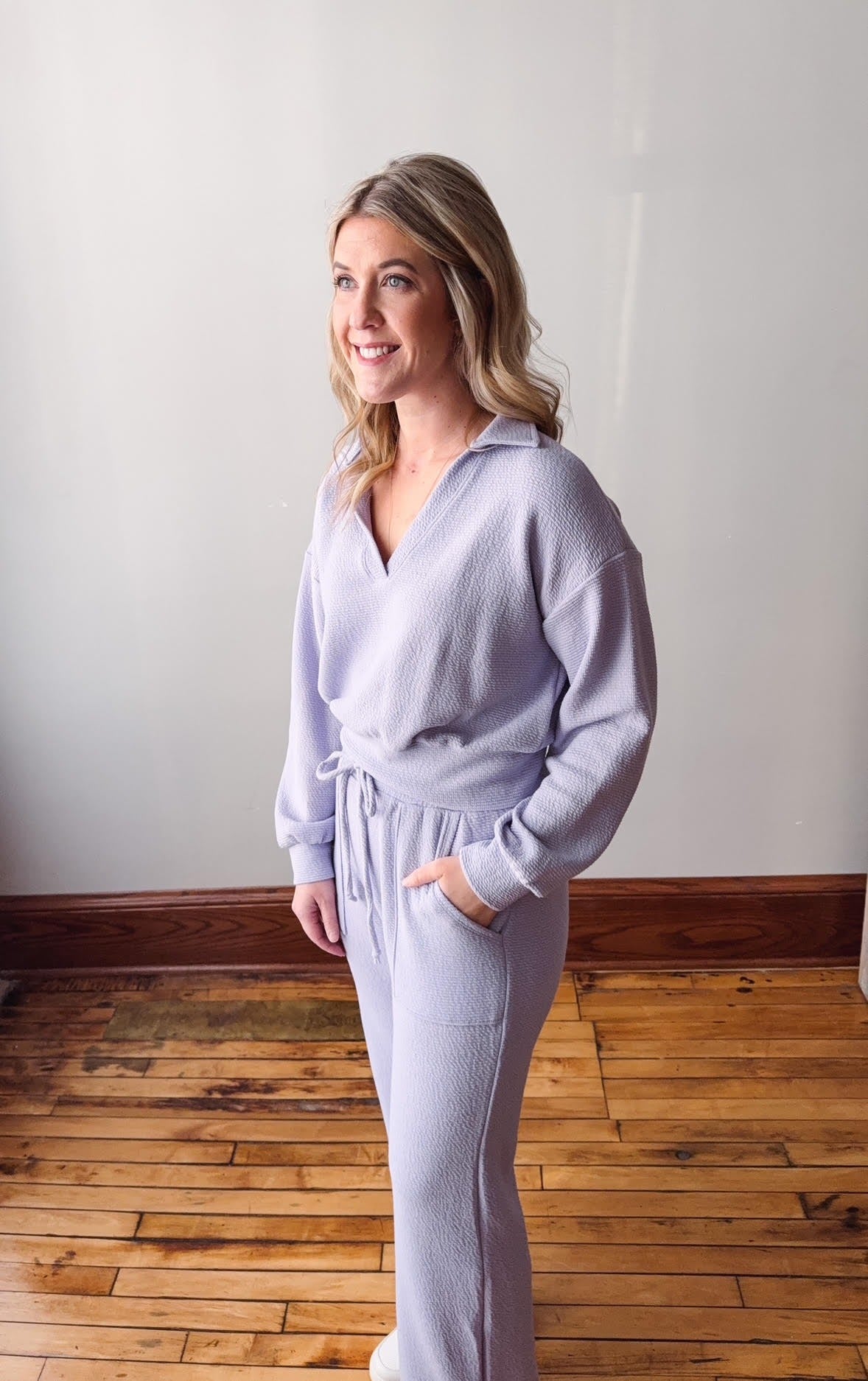 Woman wearing a light gray lounge set standing on a wooden floor with a white wall background