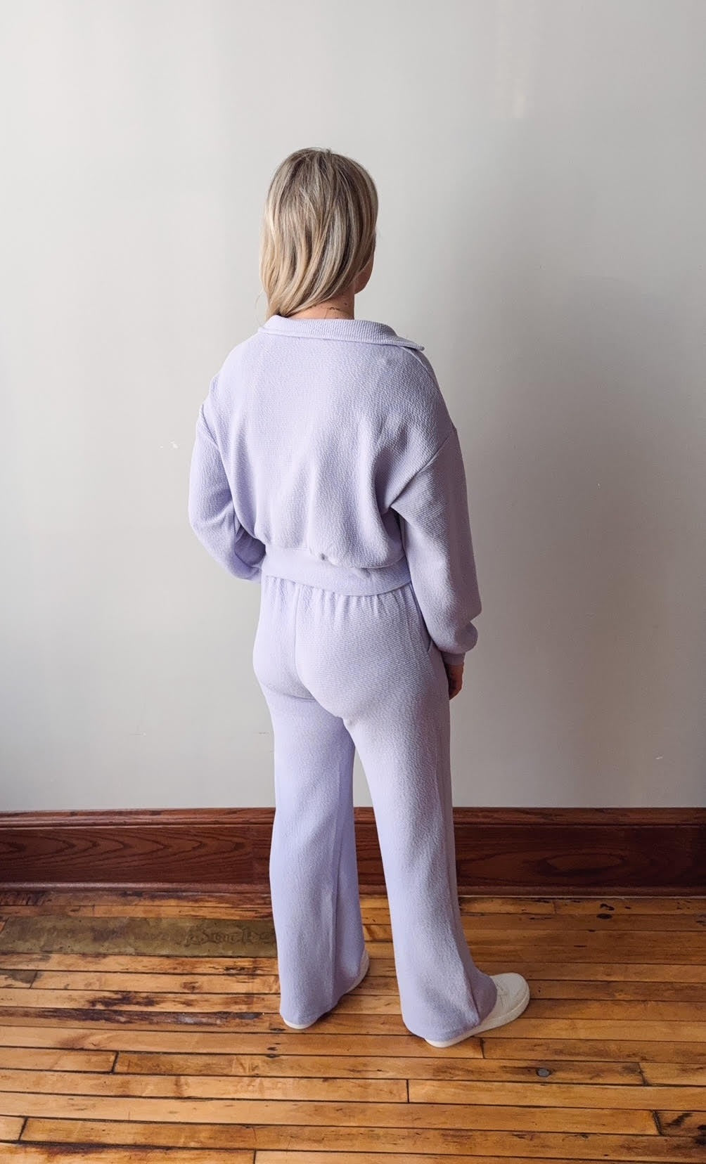 Person wearing a light purple tracksuit standing on a wooden floor against a white wall.