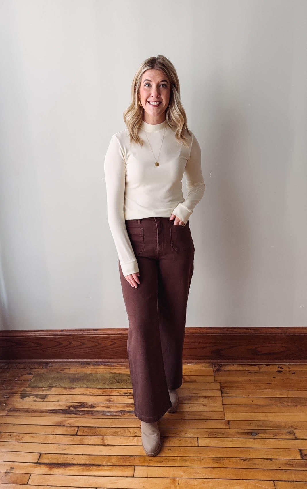 Woman wearing a white sweater and brown pants standing on a wooden floor against a plain wall.