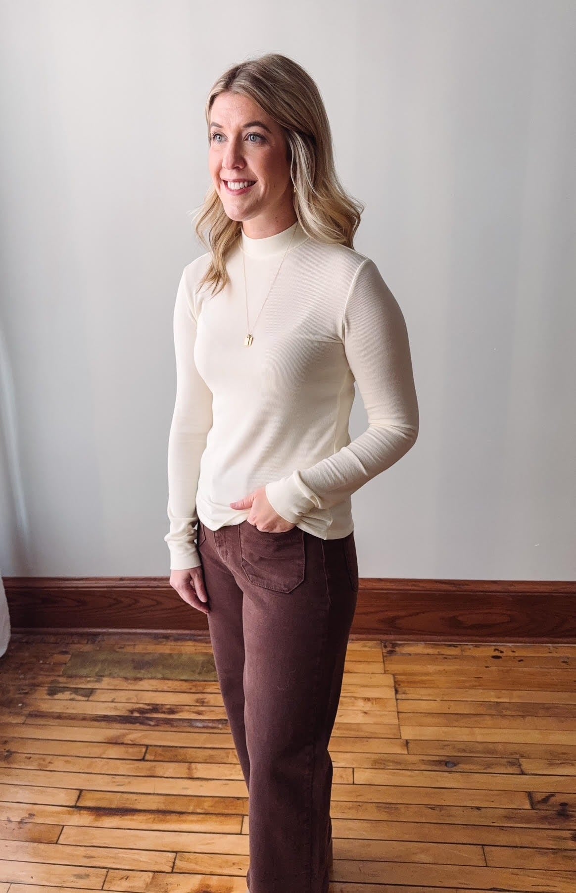 Woman wearing a white long-sleeve top and brown pants standing on a wooden floor.