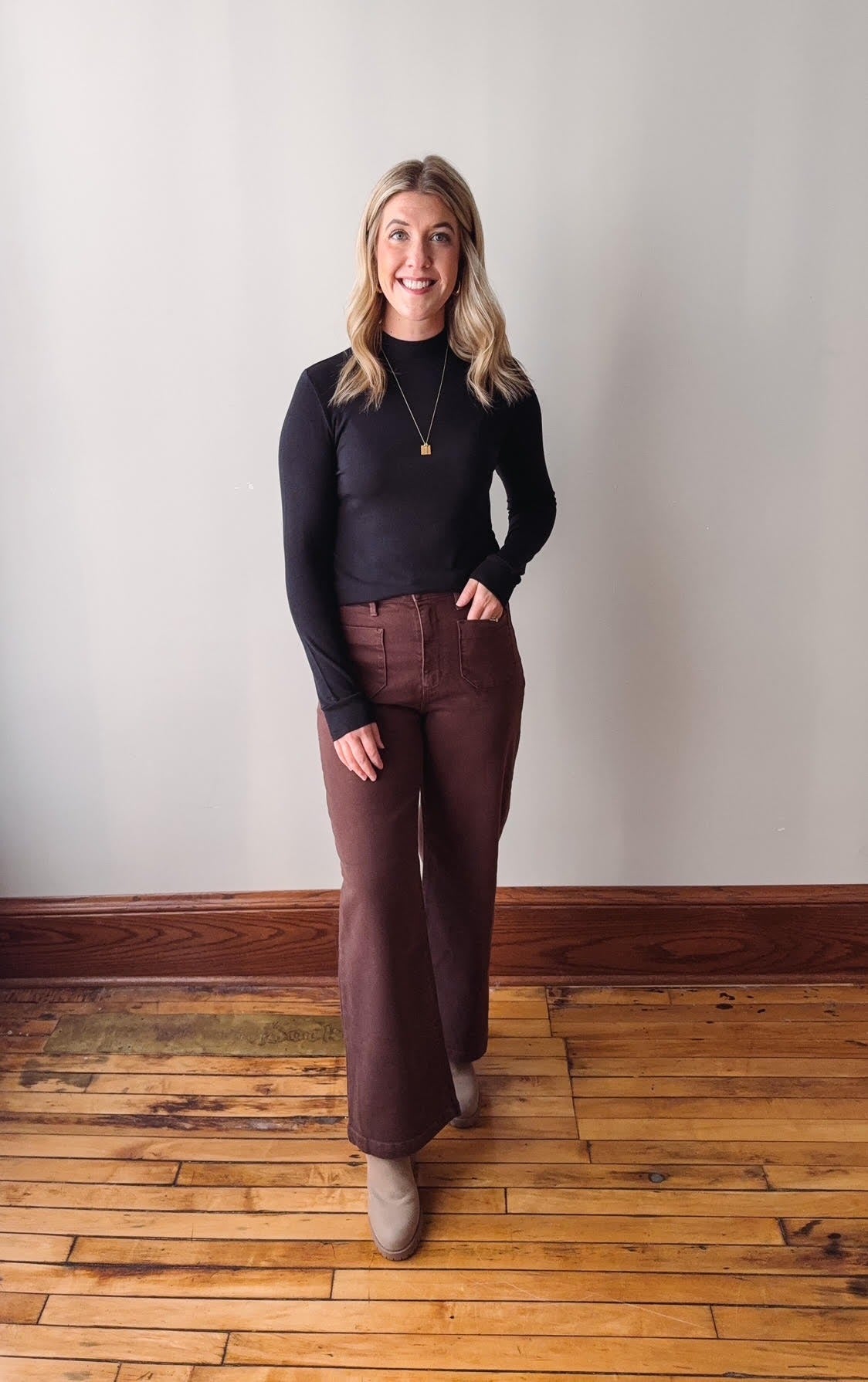 Woman wearing a black sweater and brown pants standing on a wooden floor with a white wall background