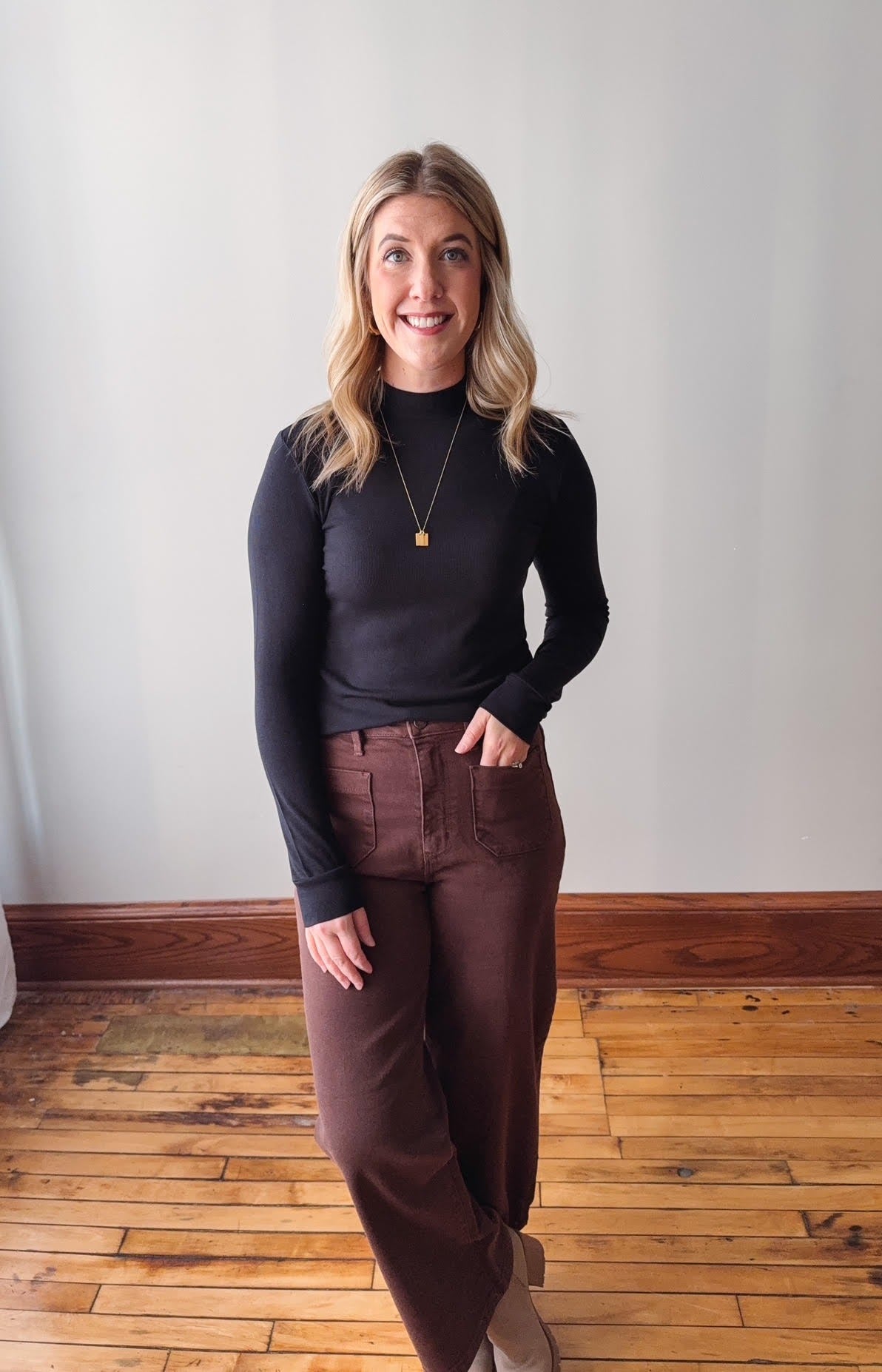 Woman wearing a black long-sleeve top and brown pants standing on a wooden floor.